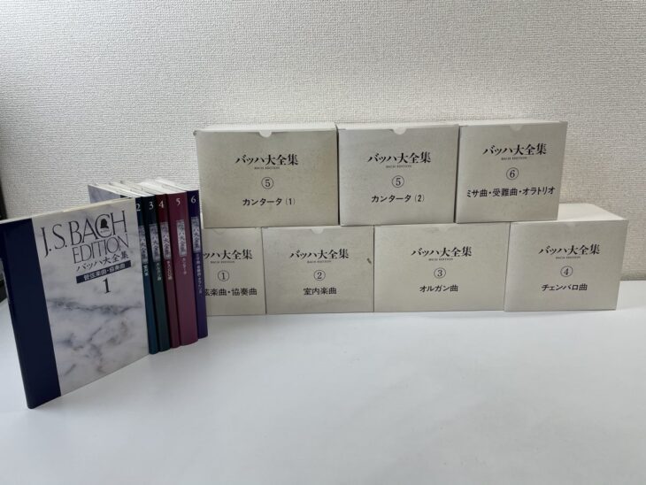 バッハ大全集の白い箱7つが2段に積まれ、左に解説書6冊と紺色の第1巻が並ぶ