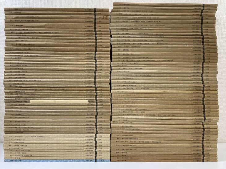 日本名跡叢刊の生成りの冊子が上下二列に約50冊ずつ積まれ、背に巻番号と各巻の作品名が入っている