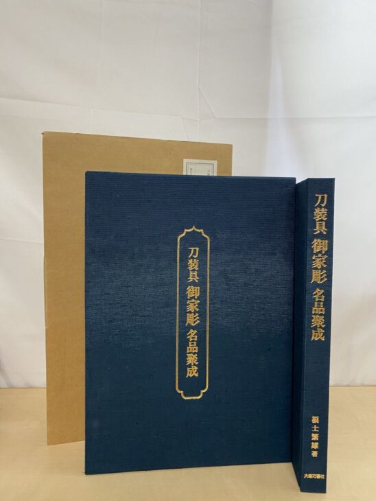 紺の布張りに金の箔押しで刀装具 御家彫 名品聚成と入った本と、後ろに厚紙の輸送函が立てて置かれている