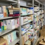 スチールの書棚が奥まで続き、『日本語文法大辞典』『ジーニアス英和辞典』が並び、手前に紐で束ねた本と段ボール箱