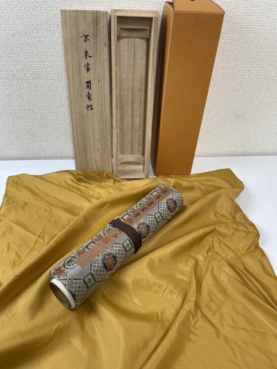 蓋に宋 米芾 蜀素帖と墨書された桐箱と橙色の外箱、黄色い布の上に置いた錦の表紙の巻子