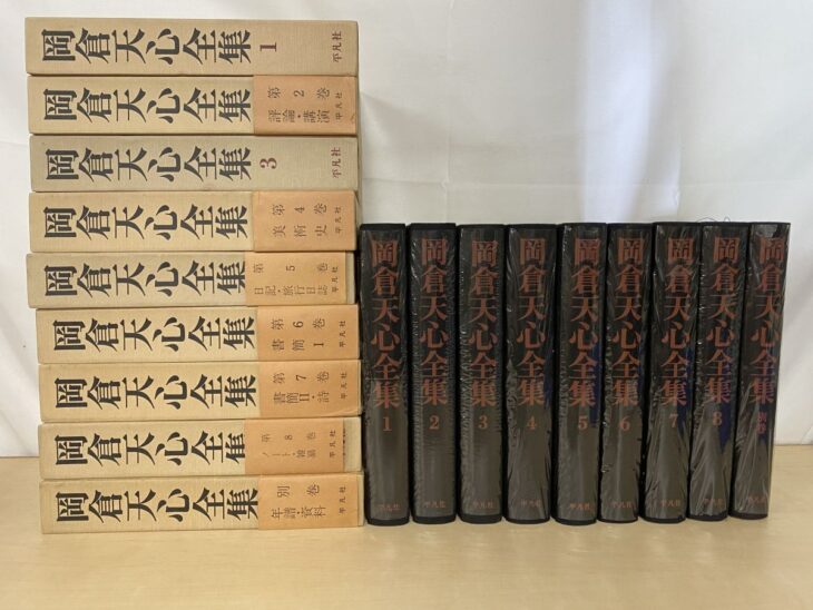 平凡社『岡倉天心全集』の生成り色の紙函9つと、透明なフィルムを掛けた黒い背に朱色の書名が並ぶ本9冊