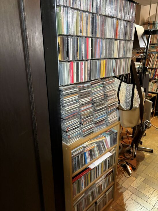 黒い棚の上段にCDが10段ほど並び、その下に積み重ねたCDと、木のラックに入ったCD