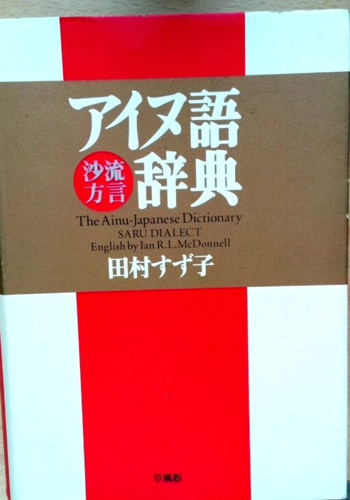 草風館『アイヌ語沙流方言辞典』の表紙。赤と白の地に茶色の帯状の枠があり、白い字の書名と「沙流方言」、英語の題、田村すず子の名前