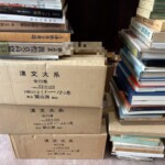 冨山房『漢文大系』全22巻の箱3つの上に『中國石窟 敦煌莫高窟』や奈良国立博物館の図録が積まれ、右に東洋文庫などの本の山が続く部屋の隅