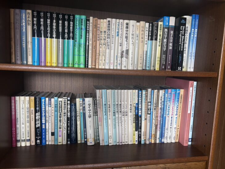 木の本棚2段。『解析入門』『線型代数入門』の基礎数学シリーズ、『フィードバック制御入門』『伝熱工学』、グライナー『量子力学概論』、『自動車力学』『タイヤ工学』『自動車技術ハンドブック』